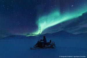 Person driving snowmobile left under a light green northern lights show in starry winter dark blue night sky in Svalbard, Arctic Norway