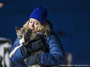 Blonde girl with dark blue beanie hat and puffer jacket holding sled dog puppy in Svalbard, Arctic Norway