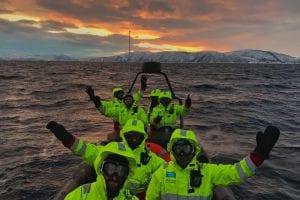 Seven people on boat safari waving their hands at the camera while at sea with mountain backdrop