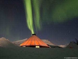 Building behind snow at Camp Tamok under light green northern lights in bright starry night sky in Lyngen, Arctic Norway