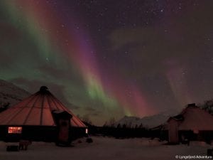 Lit up building at Camp Tamok under green and pink northern lights in starry night sky in Lyngen, Arctic Norway