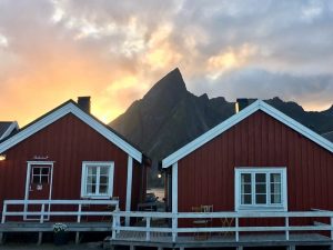 Three Eliassen Rorbuer cabin exteriors with verandas in front of sun and mountain backdrop in Hamnøy, Lofoten