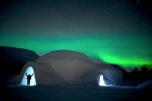 Ice domes with the silhouette of a person holding arms up at the entrance on a starry winter night with bright green northern lights in Arctic Norway