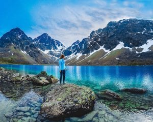 Girl dressed in blue hiking gear standing on a large flat rock looking back at clear sky, blue still water and mountains in summer in Lyngen, Arctic Norway