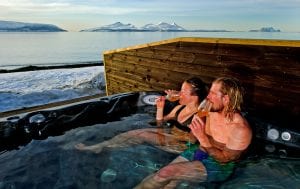 Couple sipping drinks in outdoor jacuzzi while looking out at view of sea and mountains in winter at Lyngen Experience Lodge, Arctic Norway