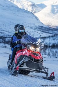 Pair in blue suits with goggles driving a red snowmobile in the mountains of Lyngen, Arctic Norway