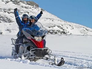 Couple on snowmobile with woman waving arms in air and man driving through snow with mountain backdrop in Lyngen, Arctic Norway