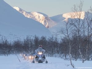 Line of snowmobiles on tour driving towards camera in bare forest with sun-kissed mountain backdrop in Lyngen, Arctic Norway